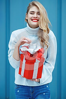 Happy pretty young woman holding a gift box over blue background.