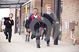 Happy primary school kids, wearing school uniforms and backpacks, running on a walkway outside their school building, front view,