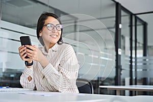 Happy professional Asian business woman using phone looking away in office.