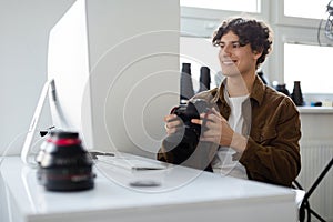 Happy professional photographer man holding camera and looking at computer monitor, sitting at workplace in studio