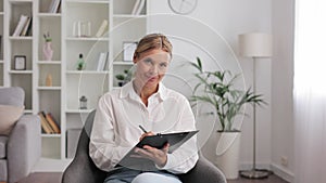 Happy professional psychologist woman sitting in chair writing on clipboard, looking and smiling at camera, working in