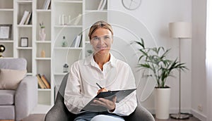 Happy professional psychologist woman sitting in chair writing on clipboard, looking and smiling at camera, working in