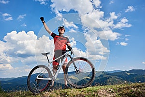 Happy professional sportsman cyclist standing with cross country bicycle on a hill, rasing hand