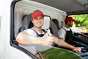 Happy professional truck driver with his assistant wearing a red cap thumb up, smiling, looking at the camera from a truck window