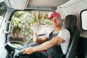 Happy professional truck driver with his assistant wearing a red cap thumb up, smiling, looking at the camera from a truck window
