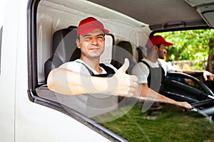 Happy professional truck driver with his assistant wearing a red cap thumb up, smiling, looking at the camera from a truck window