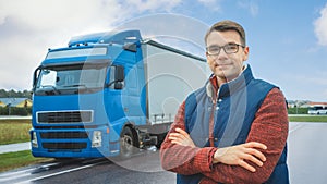 Happy Professional Young Truck Driver Crosses Arms and Smiles on Camera. Behind Him Parked Blue Long