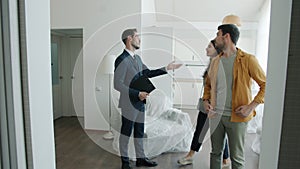 Happy real estate agent in suit showing new house to couple of buyers talking gesturing