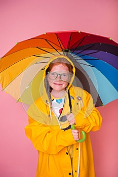 Happy red hair kid girl laughing with rainbow colored umbrella on pink background. Emotional freckled child in yellow