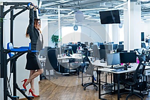 Happy red-haired business woman doing press exercises on a mounted horizontal bar in the office. A female employee in an