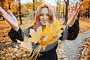 Happy red-haired woman holding yellow maple autumn leaves in fall park.