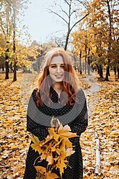 Happy red-haired woman holding yellow maple autumn leaves in fall park.