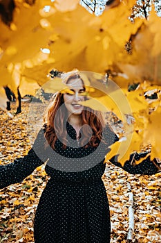 Happy red-haired woman holding yellow maple autumn leaves in fall park.