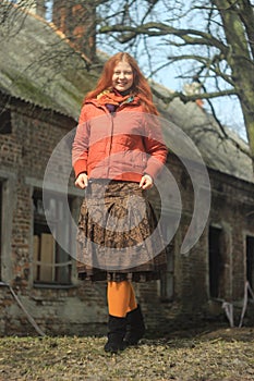 A happy red-haired young girl in a bright red jacket and pantyhose, in a skirt with floral patterns, poses against the backdrop of