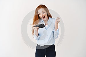 Happy redhead girl winning in mobile phone video game, saying yes and smiling, celebrating online victory, standing over