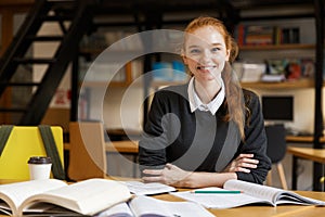 Happy redhead lady student sitting at the table with books in library