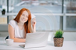 Happy redhead shopper deciding what to buy on line holding a credit card sitting