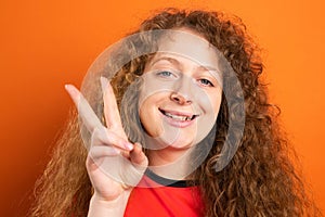 Happy redhead smiling beautiful fan football woman showing two fingers up posing on orange bavground, footbal fan