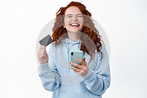 Happy redhead woman rejoicing, holding smartphone and plastic credit card, close eyes and laugh from overjoy, standing