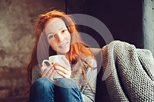 Happy redhead woman relaxing at home in cozy winter or autumn weekend with book and cup of hot tea, sitting in soft chair