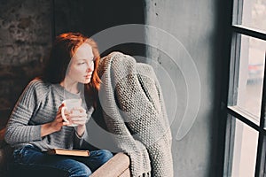 Happy redhead woman relaxing at home in cozy winter or autumn weekend with book and cup of hot tea, sitting in soft chair