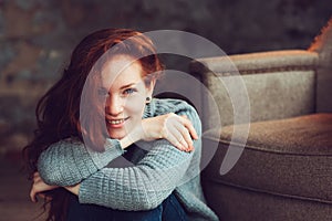 Happy redhead woman relaxing at home in cozy winter or autumn weekend with book and cup of hot tea, sitting in soft chair