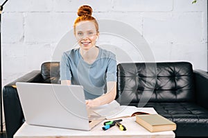 Happy redhead young business woman is working on laptop at home office, looking at camera.