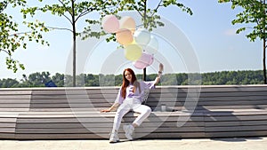 Happy redhead young woman sitting on bench with large fountain of colorful balloons outside.