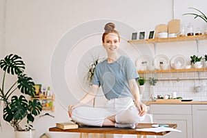 Happy redhead young woman sitting in lotus pose at the desk and looking at camera.