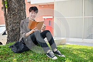 A happy, relaxed Asian male college student enjoys reading a book under the tree in the campus park