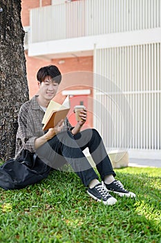A happy, relaxed Asian male college student enjoys reading a book under the tree in the campus park