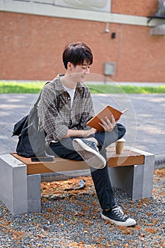 A happy, relaxed Asian male college student sits on a bench in the campus park reading a textbook