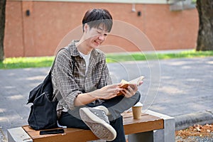 A happy, relaxed Asian male college student sits on a bench in the campus park reading a textbook