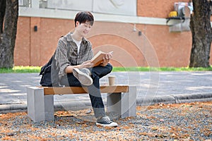 A happy, relaxed Asian male college student sits on a bench in the campus park reading a textbook