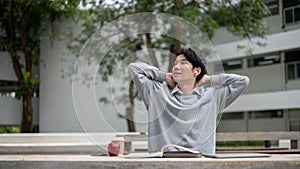 A happy and relaxed Asian male student is stretching his arms while reading books in a campus park