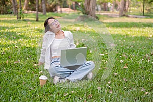 A happy, relaxed Asian woman sits on the grass in a park with her laptop, enjoying the sunlight
