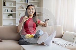 Happy relaxed asian woman watching tv and eating popcorn at home