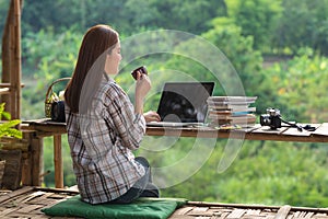 Happy relaxed asian young woman sitting drink coffee in the midst of nature with a laptop in the back of her and looking out of