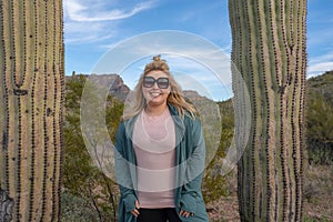 Happy, relaxed blond woman wearing sunglasses stands between two saguaro cactus cacti in Arizona, wearing casual clothing