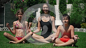 Happy relaxed Caucasian young woman little boy and girl sitting in lotus pose on green grass smiling looking at camera