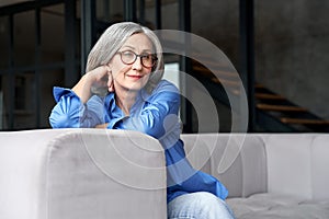 Happy relaxed mature old woman wears glasses sitting on couch at home, portrait.