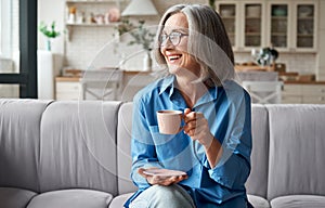 Happy relaxed mature older woman drinking coffee relaxing on sofa at home.