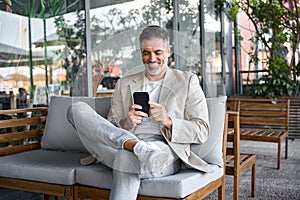Happy relaxed mid aged business man sitting in outdoor cafe using smartphone.