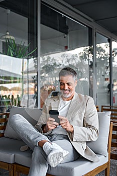 Happy relaxed mid aged business man sitting in outdoor cafe using smartphone.