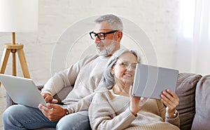 Happy relaxed old retired family couple using modern technologies laptop and digital tablet at home