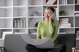 Happy and relaxed young Asian woman listening to music through her headphones on a sofa in her living room.