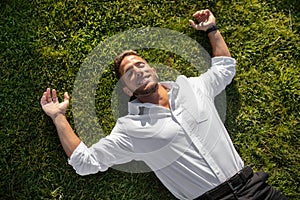 Happy relaxed young businessman in shirt lying on green grass on lawn and resting in park with closed eyes, above view