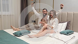 Happy relaxed young family couple lying in bed holding remote control watching tv together. Smiling millennial man and woman talki