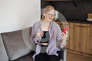 Happy relaxed young woman with glass of water sitting on couch using cell phone. 30s girl surfing useful information