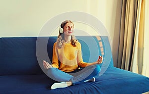 Happy relaxed young woman listening to music with wireless headphones meditating with closed eyes sitting in the lotus position on
